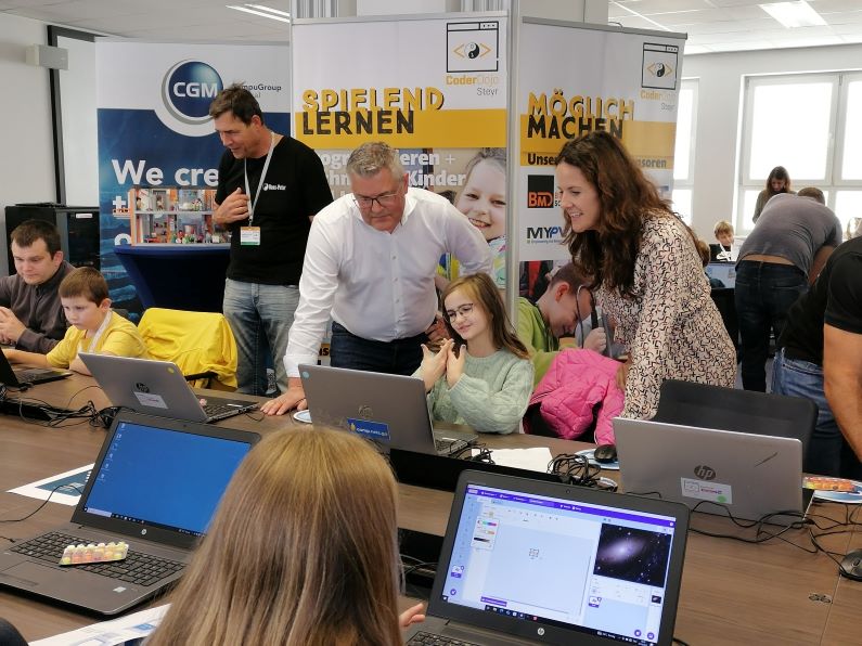 Kinder sitzen vor Computern, Erwachsene schauen ihnen über die Schultern.