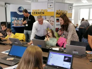 Kinder sitzen vor Computern, Erwachsene schauen ihnen über die Schultern.